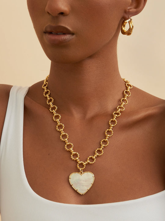 Stylish woman adorned with a chunky gold heart necklace and twisted gold hoops. Browse our collection of unique statement jewelry for any occasion.